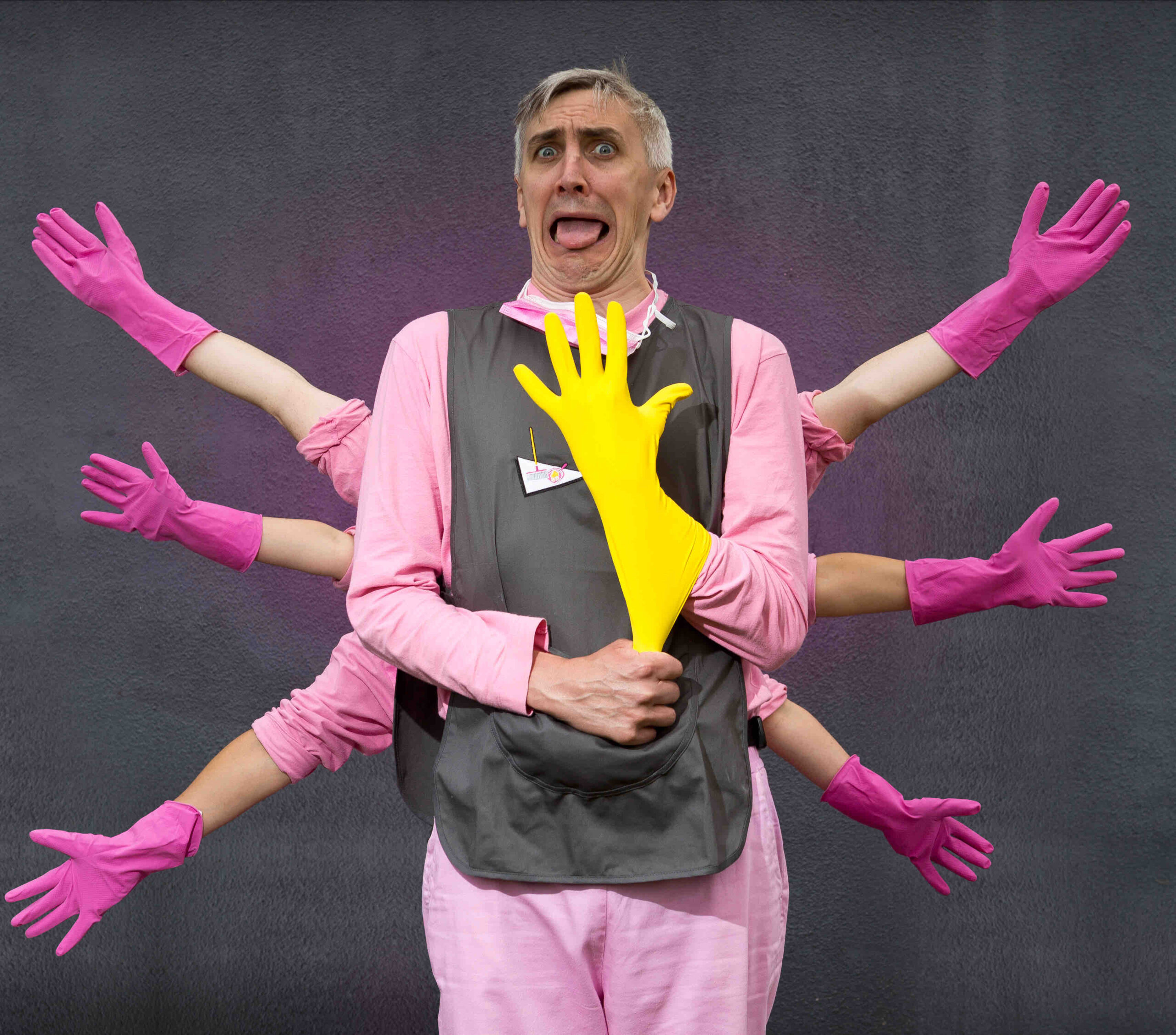 The Chief is standing against the grey painted wall and is wearing a pink t shirt and trousers with a grey tabard over the top. He is pulling a yellow rubber glove onto his hand in front of his body, and his face seems like a mixture of surprise and disgust. Behind him, the other characters’ arms fan out on either side. They are all wearing pink rubber gloves.