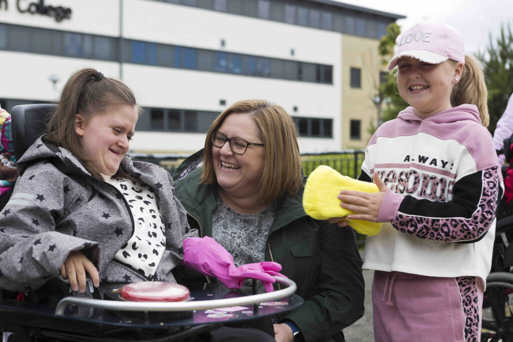 A girl sitting in a wheelchair has a big mac button on her table, and a pink glove on her hand. She appears to be smiling. Her mum is kneeling beside her and looking in her direction and seems to be laughing. Her sister is standing next to her mum, holding a big yellow sponge, and is smiling.